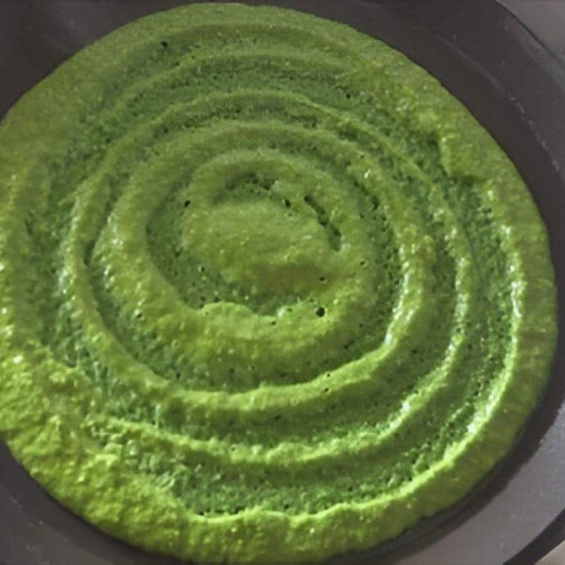 A vibrant green Indian dosa, spiraled and cooking in a dark pan, from AniKri Foods.