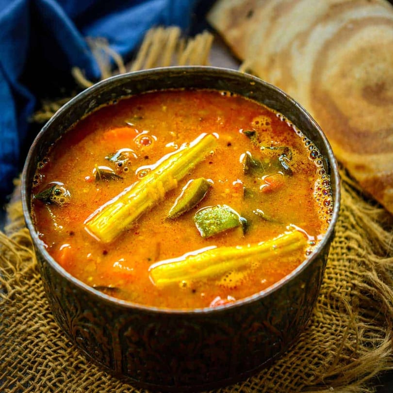 A bowl of vibrant orange Indian sambar with vegetables, served on a rustic burlap mat.