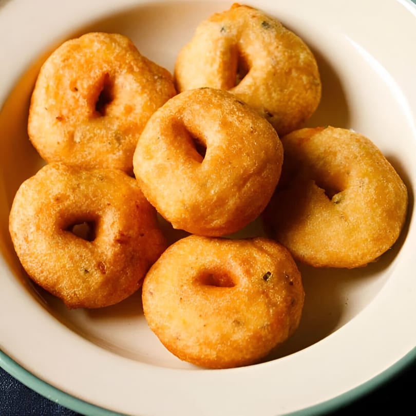 A white bowl filled with several golden-brown, ring-shaped Indian medu vada, a savory fried snack.